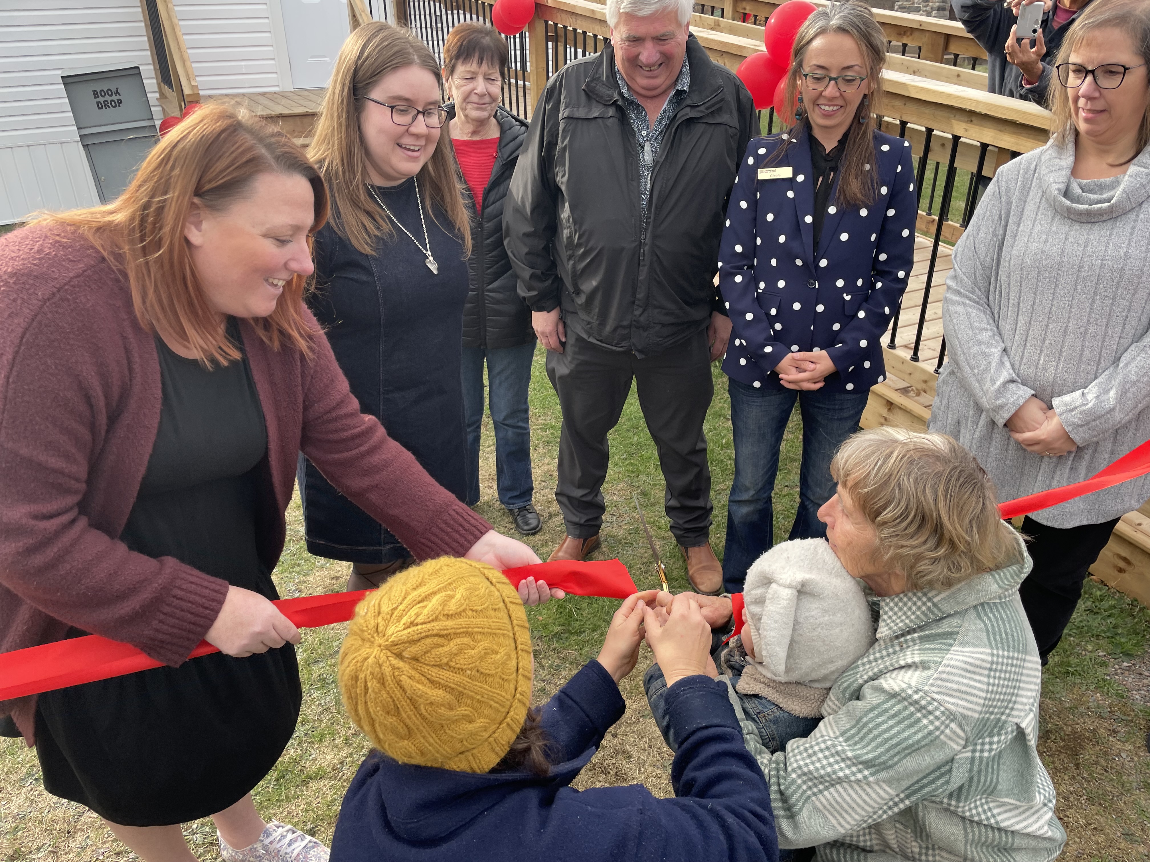 A young child is helped by three women in cutting a red ribbon while five other smiling people stand in the background.